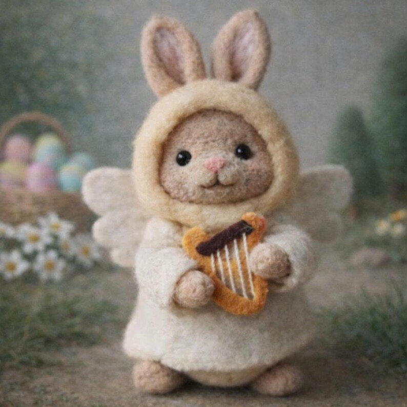 May include: A handcrafted felt bunny figurine dressed as an angel, wearing a cream-colored hooded robe and holding a small, orange harp. The bunny has large, floppy ears and small, black eyes. The background is blurred with hints of green and white.