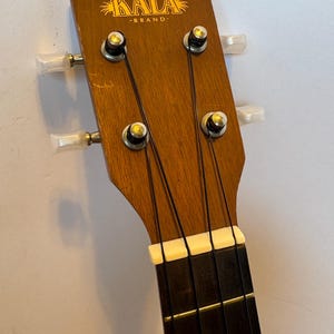 Puede incluir: Primer plano del clavijero de un ukelele. El clavijero de madera presenta el logotipo "KALA BRAND" en letras doradas. Se ven cuatro clavijas con perillas blancas, junto con las cuerdas y una cejuela blanca. El diapas&oacute;n es marr&oacute;n oscuro.