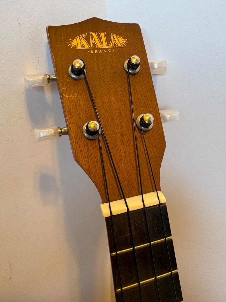 Puede incluir: Primer plano del clavijero de un ukelele. El clavijero de madera presenta el logotipo "KALA BRAND" en letras doradas. Se ven cuatro clavijas con perillas blancas, junto con las cuerdas y una cejuela blanca. El diapas&oacute;n es marr&oacute;n oscuro.
