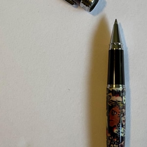 May include: A rollerball pen with a decorative design. The pen has a silver clip and tip, with a black barrel and a colorful pattern featuring faces and figures. The cap is detached, lying next to the pen.