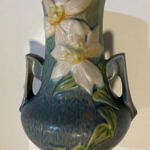 Puede incluir: Un jarrón de cerámica azul con dos asas, con un diseño floral de flores blancas y amarillas con hojas verdes. El jarrón tiene una superficie texturizada y un borde ligeramente ensanchado, mostrando una estética artesanal.