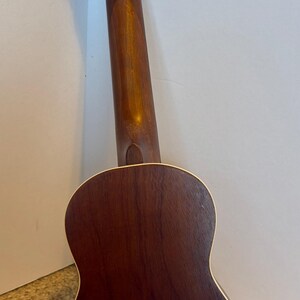 Puede incluir: Vista trasera de un ukelele, que muestra su cuerpo y m&aacute;stil de madera marr&oacute;n oscuro. El instrumento tiene cuatro clavijas de afinaci&oacute;n con perillas blancas. El cuerpo tiene una forma redondeada con un borde de color claro.