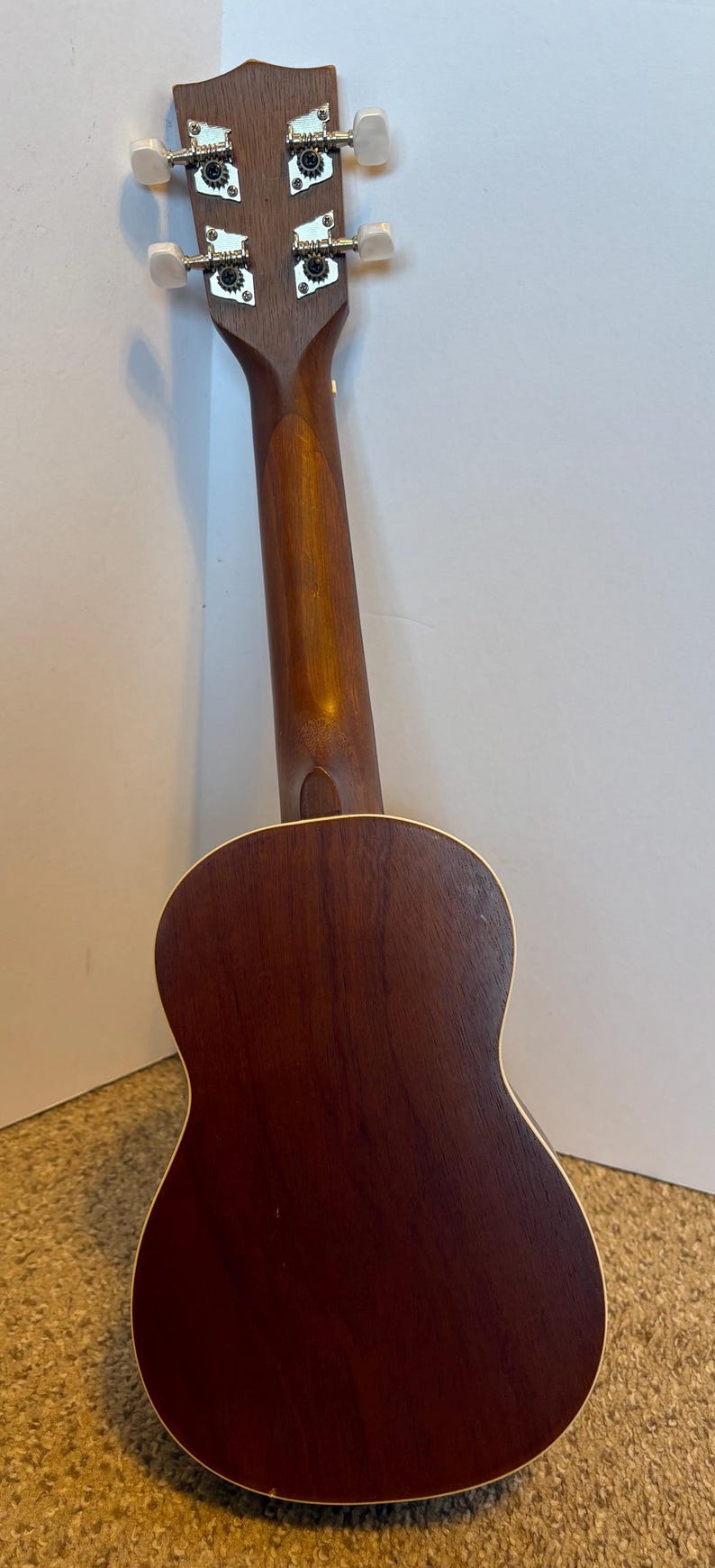 Puede incluir: Vista trasera de un ukelele, que muestra su cuerpo y m&aacute;stil de madera marr&oacute;n oscuro. El instrumento tiene cuatro clavijas de afinaci&oacute;n con perillas blancas. El cuerpo tiene una forma redondeada con un borde de color claro.