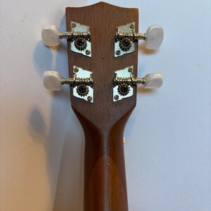 Puede incluir: Primer plano del clavijero de un ukelele, mostrando las clavijas y la construcci&oacute;n de madera. El clavijero es de color marr&oacute;n oscuro, con cuatro clavijas plateadas con perillas blancas. El m&aacute;stil del ukelele tambi&eacute;n es visible, en un marr&oacute;n m&aacute;s claro.