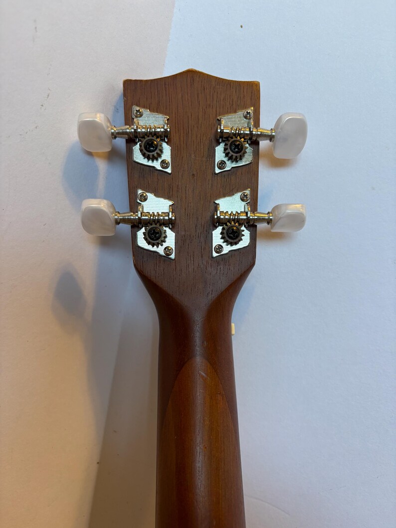 Puede incluir: Primer plano del clavijero de un ukelele, mostrando las clavijas y la construcci&oacute;n de madera. El clavijero es de color marr&oacute;n oscuro, con cuatro clavijas plateadas con perillas blancas. El m&aacute;stil del ukelele tambi&eacute;n es visible, en un marr&oacute;n m&aacute;s claro.