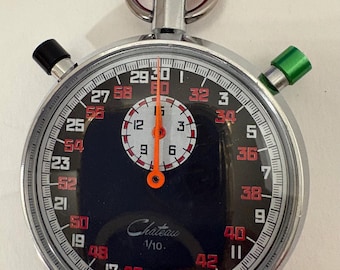 Puede incluir: Un cronómetro Chateau vintage con esfera negra, caja plateada y detalles en rojo y verde. El reloj presenta una esfera central con números y agujas, y una corona roja. El texto "Chateau 1/10" es visible.