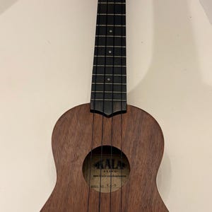Puede incluir: Un ukelele marr&oacute;n de la marca Kala con un agujero de sonido redondo y cuerdas negras. El ukelele tiene un acabado de madera oscura y un diapas&oacute;n negro. El clavijero tiene clavijas de afinaci&oacute;n.