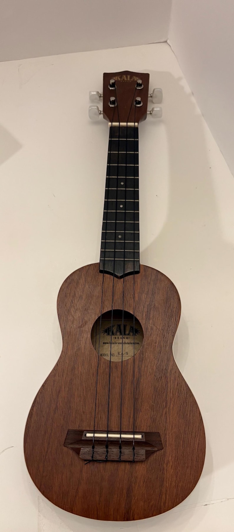 Puede incluir: Un ukelele marr&oacute;n de la marca Kala con un agujero de sonido redondo y cuerdas negras. El ukelele tiene un acabado de madera oscura y un diapas&oacute;n negro. El clavijero tiene clavijas de afinaci&oacute;n.