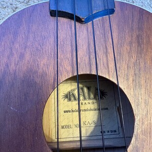 Puede incluir: Primer plano de un ukelele marr&oacute;n con cuerdas negras. La boca revela una etiqueta con la marca "KALA" y el modelo "KA-S". El cuerpo de madera del instrumento muestra una veta natural.