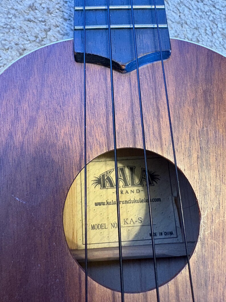 Puede incluir: Primer plano de un ukelele marr&oacute;n con cuerdas negras. La boca revela una etiqueta con la marca "KALA" y el modelo "KA-S". El cuerpo de madera del instrumento muestra una veta natural.