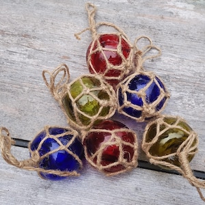 May include: A collection of six glass fishing floats in various colors, including red, green, and blue. Each float is encased in a knotted jute net and has a jute loop for hanging. The floats are arranged on a weathered wooden surface.