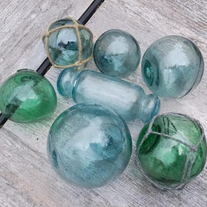 May include: A collection of vintage glass fishing floats in various shades of blue and green. Some floats are spherical, while others are cylindrical. Several are encased in netting.