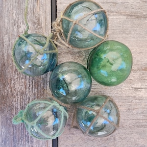 May include: Five glass fishing floats in shades of green and blue, some with netting and rope detailing. The spheres vary in size and are arranged on a weathered wooden surface, evoking a nautical theme.