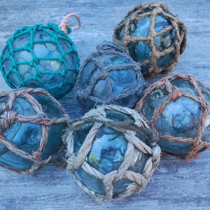 May include: A collection of six glass fishing floats in various shades of blue and green. Each float is encased in a net made of rope, with some featuring orange and tan accents. The floats are arranged on a weathered, light blue surface.