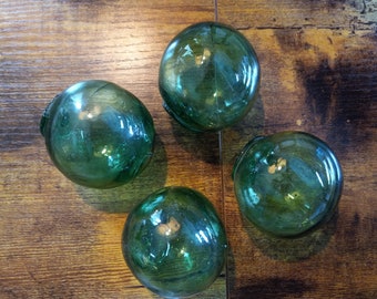 May include: Four green glass fishing floats on a wooden surface. The floats are round and have a smooth, shiny finish.