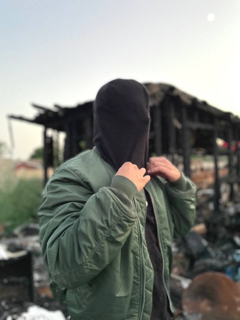 May include: A person wearing a green bomber jacket with a black hooded sweatshirt pulled over their head. The person is standing in front of a burned-out building.