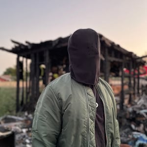 May include: A person wearing a black balaclava and a green bomber jacket stands in front of a burned-out building.