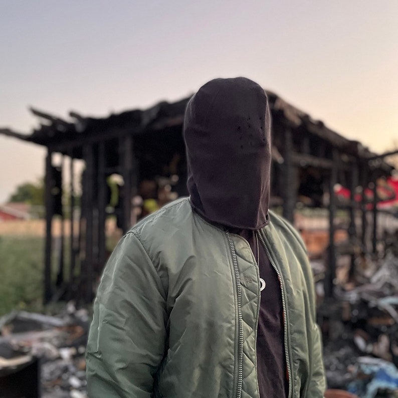 May include: A person wearing a black balaclava and a green bomber jacket stands in front of a burned-out building.