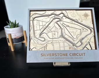 May include: A wooden model of the Silverstone Circuit race track, with a grey frame and a silver plaque that reads "Silverstone Circuit, Towcester, Northamptonshire, England".