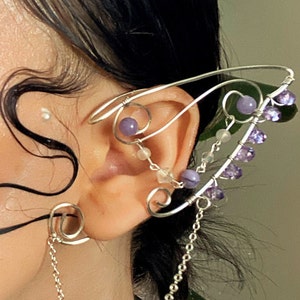 Puede incluir: Un pendiente de oreja de plata con una cadena delicada y cuentas moradas. El pendiente está diseñado para envolver la oreja y tiene una cadena colgante que llega al cuello.