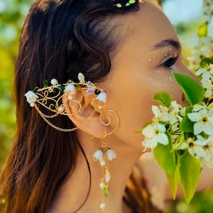 May include: A gold ear cuff with white flower accents and a dangling chain with white flowers and green beads. The ear cuff is worn by a person with long dark hair.