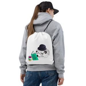 May include: White drawstring bag with a cartoon hedgehog wearing a black hat and glasses, reading a book that says "Pin Your Dreams High". A mug with a purple liquid is next to the hedgehog.