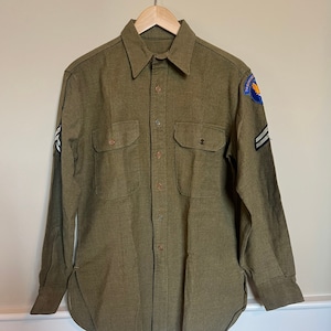 May include: A green long-sleeved shirt with two pockets and a button-up front. The shirt has a patch on the right sleeve with the text "Training" and a graphic of a plane. There are two stripes on the left sleeve.