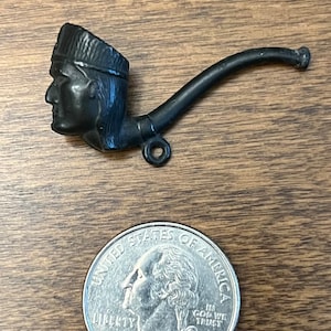 May include: A small, dark-colored pipe with a Native American profile carved into the bowl. The pipe is placed next to a United States quarter for size comparison. The pipe has a curved stem and a small loop.