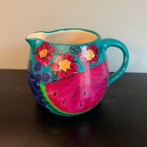 May include: A turquoise ceramic pitcher with a hand-painted design of watermelon, grapes, and flowers. The pitcher has a rounded body and a handle.