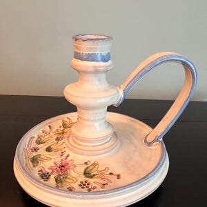 May include: A ceramic candle holder with a cream-colored base and a central socket. The piece features a blue painted rim and a curved handle. The base is decorated with a hand-painted floral pattern in shades of pink, green, and yellow.