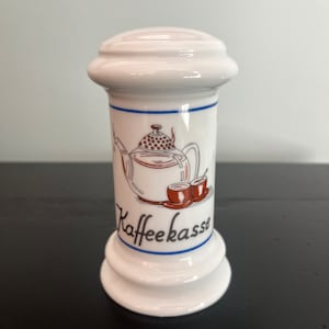 May include: White ceramic coffee container with a rounded top and base. The container features a brown and blue illustration of a teapot and cups, with the word "Kaffeekasse" in script. A blue line accents the top and bottom.