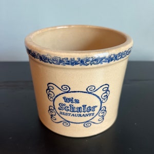May include: A beige ceramic crock with a blue and white floral design around the top and a blue and white logo that reads "Win Schuler Restaurants".