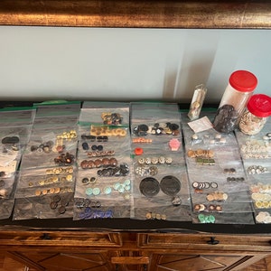 May include: A collection of various buttons in different colors, shapes, and sizes, organized in plastic bags on a wooden surface. The buttons are arranged in rows and columns, with some bags containing more than one type of button. There are also two glass jars with buttons in them.