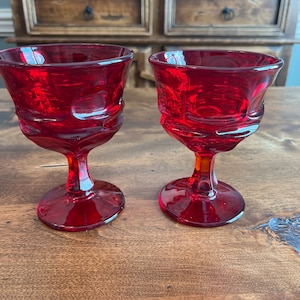 May include: Two red glass goblets with a textured design. The goblets are on a wooden surface.