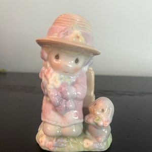 May include: A porcelain figurine of a girl wearing a pink dress and a brown hat, holding flowers. She is standing next to a small dog wearing a pink dress. The figurine is on a green base with flowers.