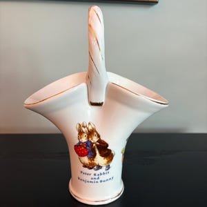 May include: White ceramic basket with a handle, featuring gold trim and a Peter Rabbit and Benjamin Bunny illustration. The basket has a flared shape and is likely for decorative use.