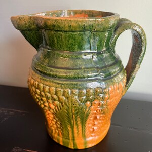 May include: A green and brown glazed ceramic pitcher with a textured design. The pitcher has a wide mouth and a handle.