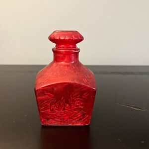 May include: A red glass bottle with a textured surface and a decorative stopper. The bottle is square shaped and has a slightly rounded bottom.