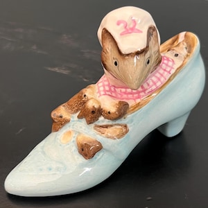 May include: A blue ceramic shoe with a pink and white gingham-patterned mouse and three baby mice inside. The mouse is wearing a white hat with a pink number "2" on it.