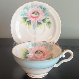 May include: A vintage teacup and saucer set with a delicate floral design. The cup is white with a light blue band and a pink flower with green leaves. The saucer is white with a pink flower and green leaves.
