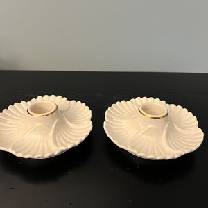 May include: Two white ceramic candle holders with a gold rim. The holders are shaped like a shell and have a scalloped edge.