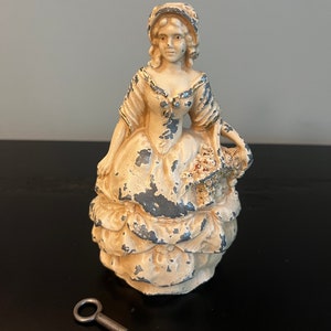May include: A vintage white cast iron figurine of a woman in a dress holding a basket of flowers. The figurine has a keyhole on the back and comes with a small metal key.