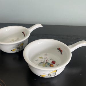 May include: Two white ceramic ramekins with handles. The ramekins have a floral design with butterflies and strawberries.