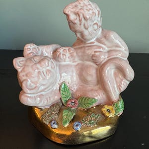 May include: A pink ceramic figurine depicting a cherub reclining on a lion. The base is gold with floral and berry accents. The figurine is a decorative item.