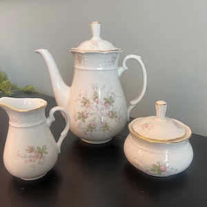 May include: A white porcelain tea set with a floral pattern. The set includes a teapot, creamer, and sugar bowl. The teapot has a gold trim around the lid and spout. The creamer and sugar bowl have gold trim around the rim.