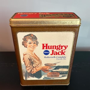 May include: Vintage Hungry Jack Buttermilk Complete Pancake Mix tin. The rectangular tin has a gold-colored top and sides with a white label featuring a woman making pancakes. The Hungry Jack logo is in red and the Pillsbury logo is in blue.