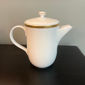 May include: White ceramic coffee pot with a lid and a curved handle. The pot has a gold band around the rim and a gold accent on the lid's knob. The spout is angled for easy pouring. The pot is on a black surface.