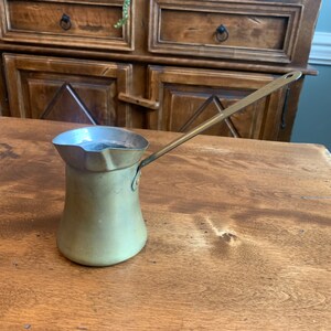 May include: A brass Turkish coffee pot with a long handle. The pot is on a wooden table.