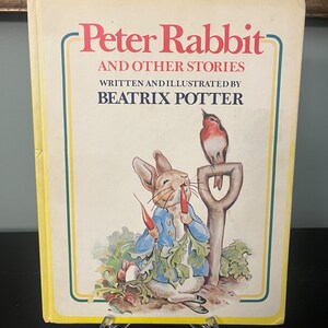 May include: A vintage children's book cover featuring Peter Rabbit, a rabbit wearing a blue jacket, eating a carrot. The book title is "Peter Rabbit and Other Stories" written and illustrated by Beatrix Potter. A robin is perched on a garden tool.