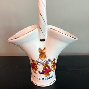 May include: White ceramic basket with a handle, featuring a gold trim and a painted illustration of Mrs. Rabbit and her children. The basket has a flared shape and the words "Mrs. Rabbit" are written in blue.